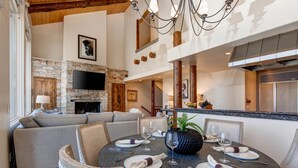 A dining area with a round table set for six, a stone fireplace, and a modern kitchen in the background. - 4 Bdr/4.5Bath-Steps to lifts and Stein Eriksen (Park City)