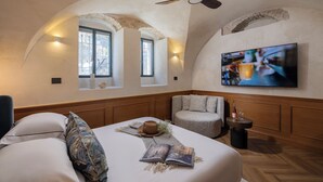 A cozy room with a bed, a round armchair, a small table with a bottle and glasses, and a TV on the wall. -  Villa Albi - Machne Yehuda Hotel (Jerusalem)