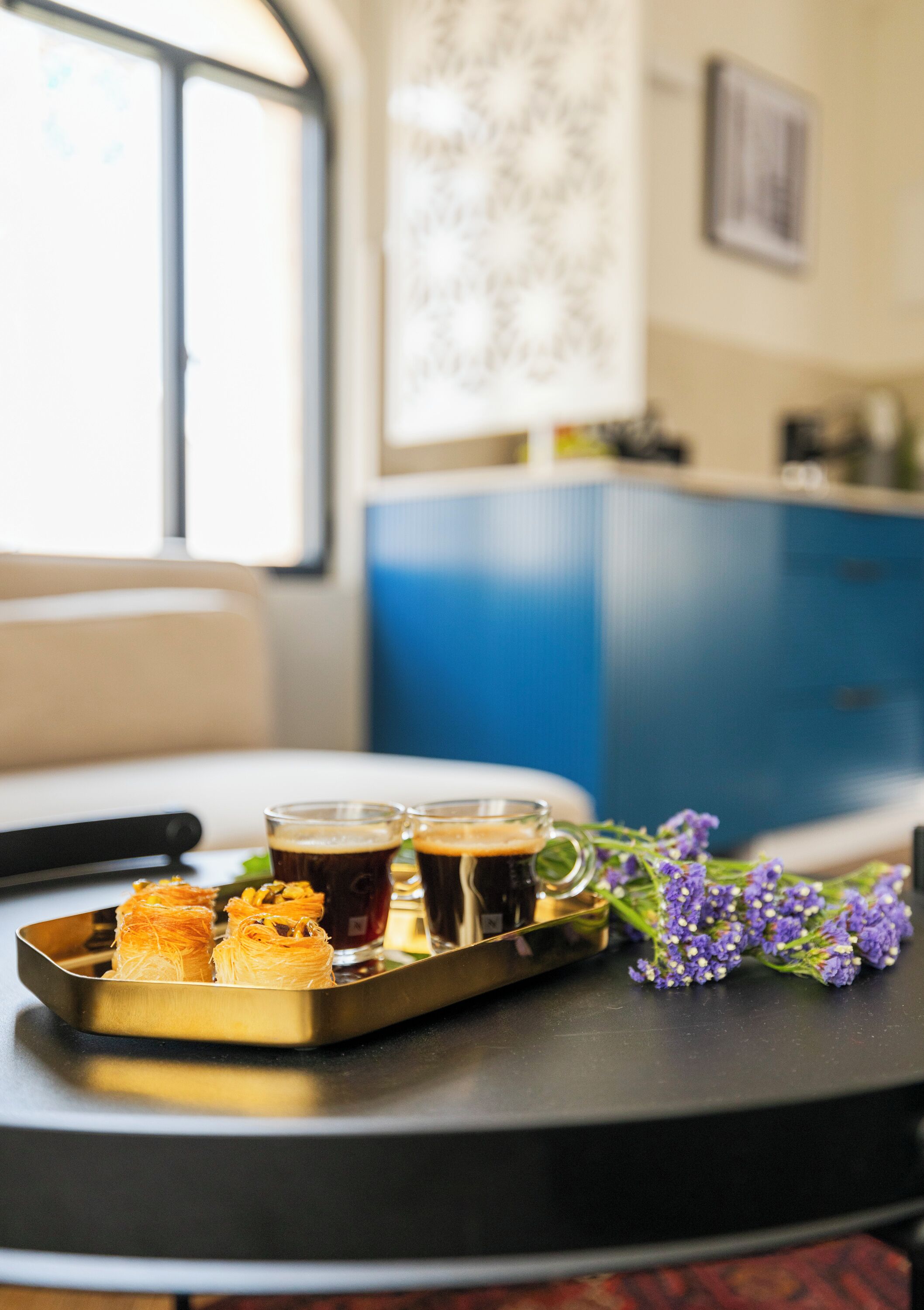 A tray with pastries and drinks on a table, with a window and a patterned wall in the background.