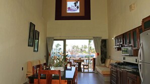 A spacious kitchen with wooden cabinets, a dining area with a table and chairs, and a large window with a view of palm trees. - VILLAS DULCE SUEÑOS (Rincón de Guayabitos)