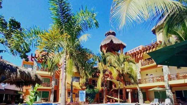 A pool area with a thatched-roof structure, palm trees, and a building with balconies. - VILLAS DULCE SUEÑOS (Rincón de Guayabitos)