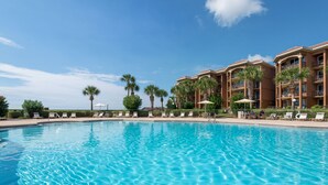 Una piscina azul cristalina con sillas de descanso y palmeras, rodeada por un edificio de hotel de varios pisos.