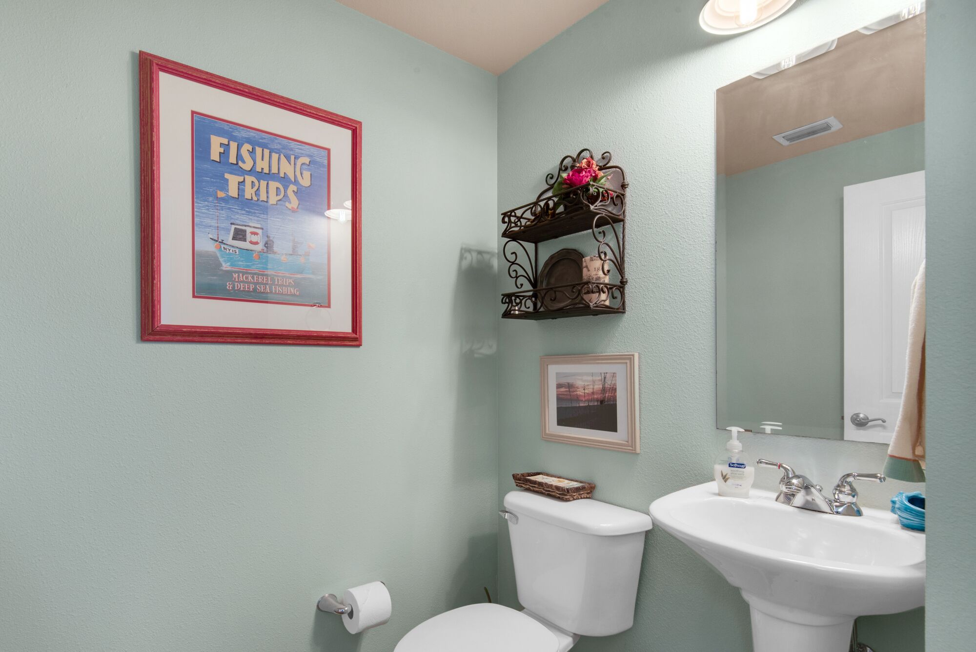 A bathroom with a toilet, sink, framed poster, and decorative shelf.
