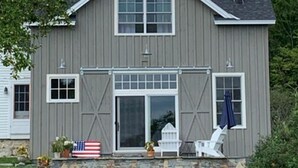 A two-story house with a stone foundation, a front porch with a flag, and a chimney. - Remodeled Barn in Champlain Valley, Essex, NY (Essex)