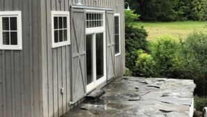 A gray wooden house with a stone-paved porch and a small garden area. - Remodeled Barn in Champlain Valley, Essex, NY (Essex)