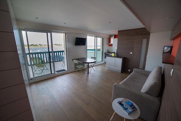 A modern living room with a sofa, a dining table, and a balcony with a view of the sea.