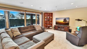 A modern living room with a sectional sofa, a flat-screen TV, a bookshelf, and a large window offering a view of the outdoors.