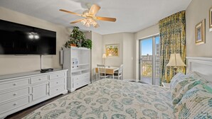 Une chambre avec un grand lit, un ventilateur de plafond, une télévision, un bureau avec une chaise et une fenêtre avec des rideaux.
