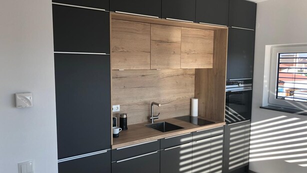 A modern kitchen with black cabinetry, a wooden accent wall, a sink, and a window with blinds. - Kruft-apps (Kruft)