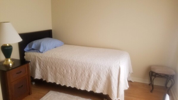 Una cama individual con una colcha blanca, una almohada azul y una mesita de noche con una lámpara.