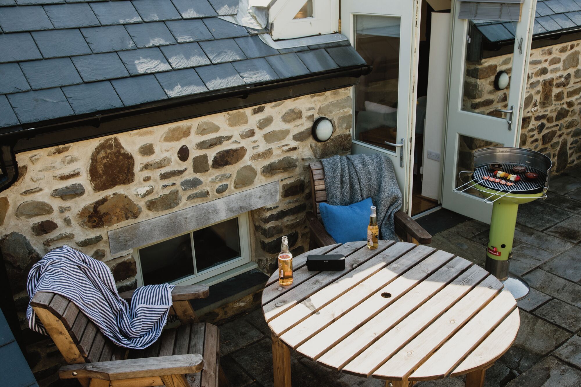 A stone building with a tiled roof, a round wooden table with a bottle and a remote control, and a grill outside.