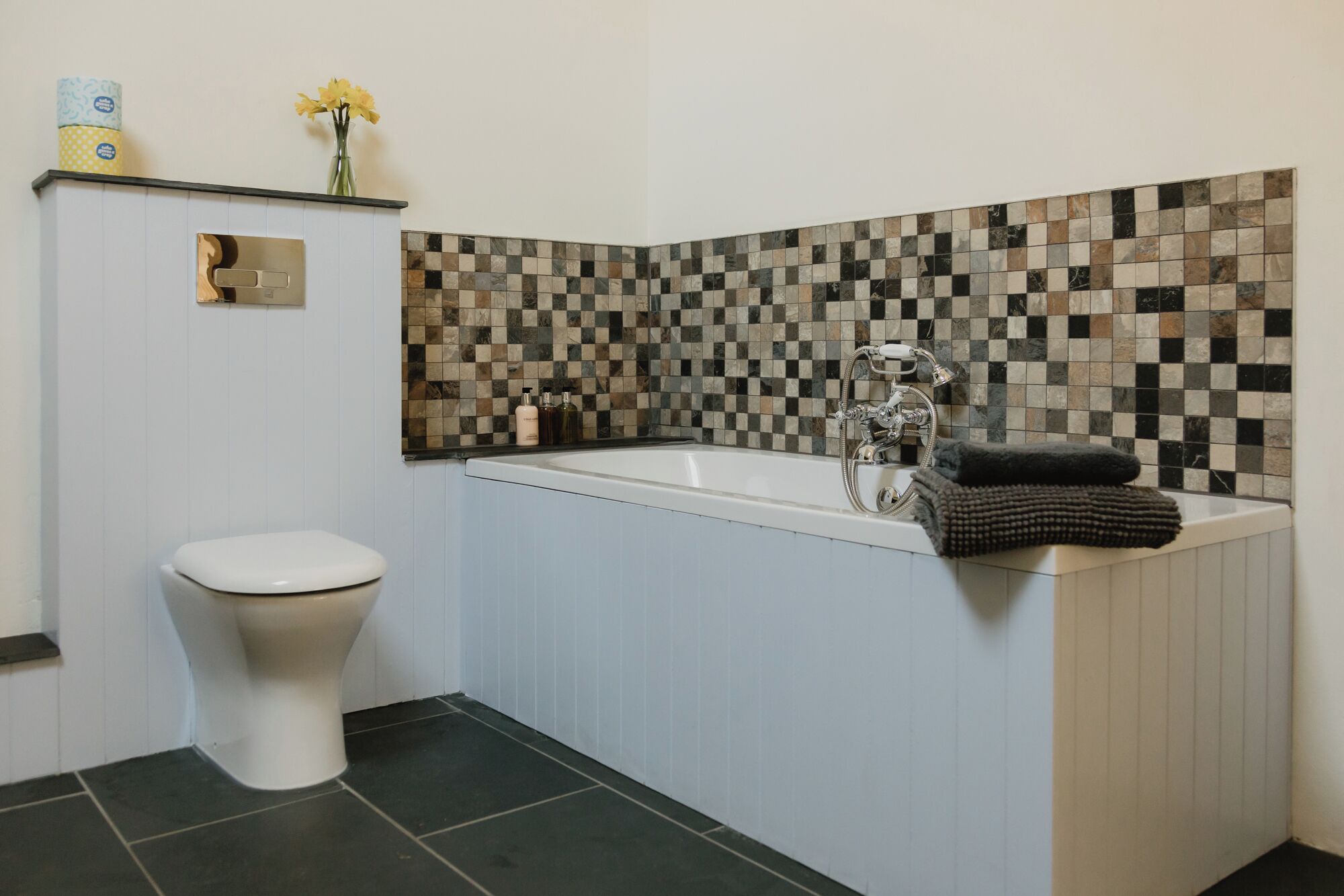 A bathroom with a toilet, a bathtub with a shower, and a mosaic tile wall.