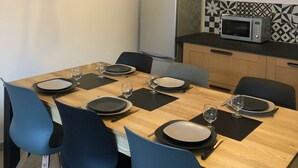 Un comedor con una mesa de madera dispuesta para cuatro personas, sillas azules, un reloj de pared y una cocina con microondas y refrigerador.