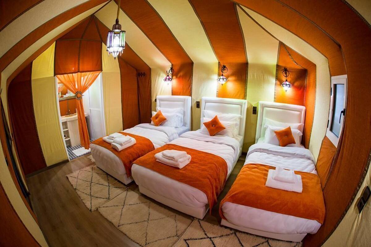 Une chambre avec trois lits, chacun doté d’une tête de lit blanche et d’une literie orange. On y trouve des lampes fixées au mur et une suspension.