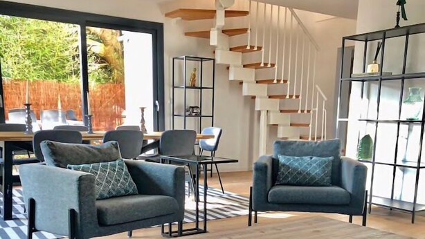 A modern living room with a wooden coffee table, two grey sofas with patterned cushions, a dining area with a table and chairs, and a staircase with a white railing.