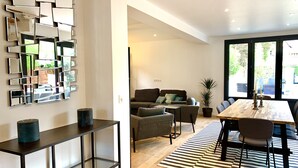 A modern living room with a sofa, coffee table, and dining table. There is a large mirror on the wall and a potted plant.