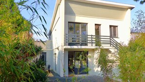 A two-story modern house with a balcony, surrounded by lush greenery.