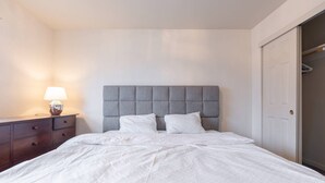 Una cama bien tendida con un cabecero tapizado en gris, una mesita de noche con una lámpara y una puerta cerrada a la derecha.