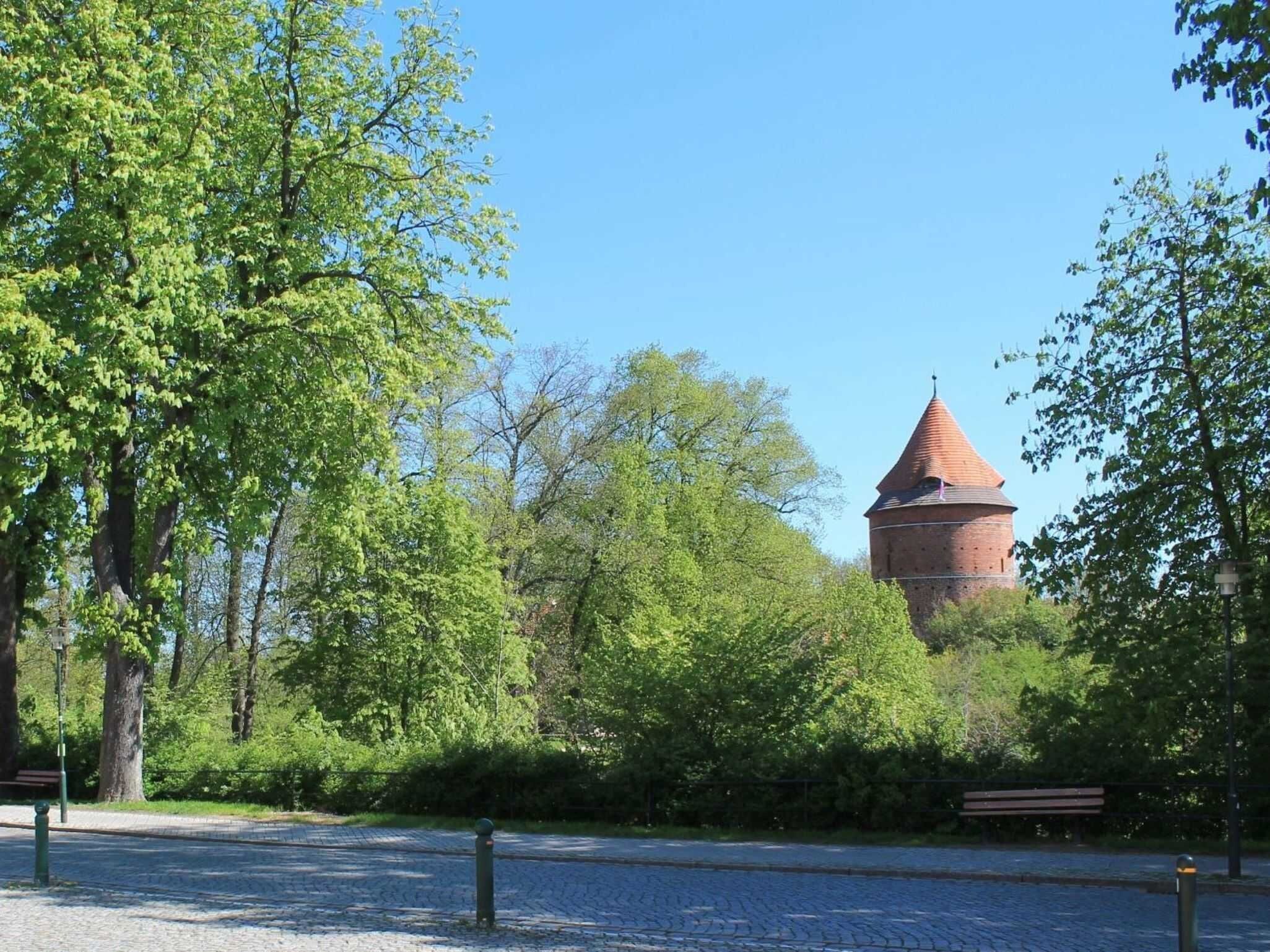 Ein Backsteinturm mit einem spitzen Dach, umgeben von üppigen grünen Bäumen und einem Kopfsteinpflasterweg.