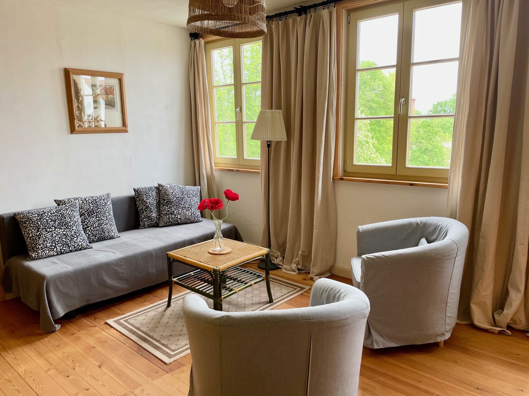 Una sala de estar con un sofá, dos sillones, una mesita con un jarrón de flores, una lámpara de pie y una ventana con cortinas.