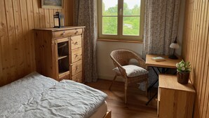 Une pièce aux murs en bois, un lit recouvert d’une couverture à motifs, une chaise en osier, un bureau avec une lampe et une fenêtre avec des rideaux.