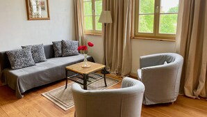 Un salon comprenant un canapé, deux fauteuils, une petite table sur laquelle est posé un vase de fleurs, un lampadaire et une fenêtre avec des rideaux.