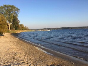A sandy beach with a calm lake, trees, and a distant dock with boats.