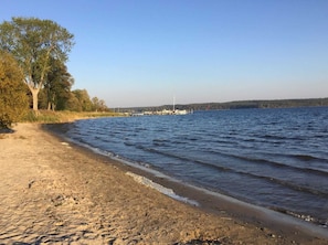 A sandy beach with a calm lake, trees, and a distant dock with boats.