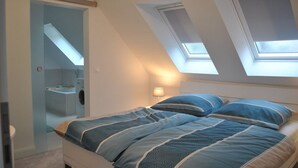 Une chambre avec une lucarne, un lit avec une literie bleue, et une salle de bain visible, équipée de toilettes et d’un lavabo.