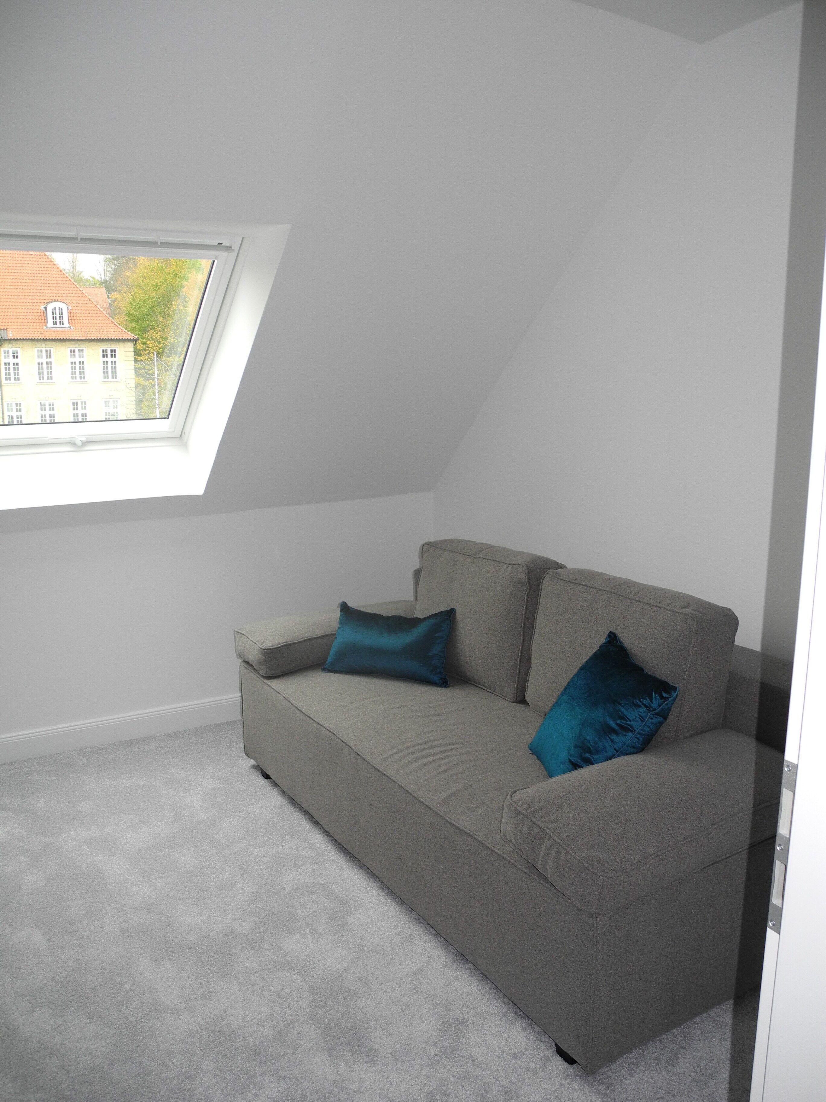 A room with a grey sofa, two cushions, a skylight, and a view of a building outside.