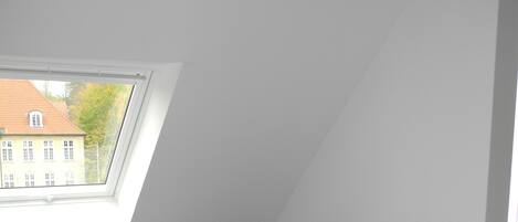 A room with a grey sofa, two cushions, a skylight, and a view of a building outside.