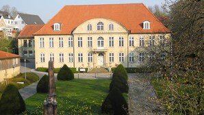 Un grand bâtiment historique, doté d’un toit recouvert de tuiles rouges et de fenêtres disposées de manière symétrique, entouré de jardins bien entretenus et surmonté d’un monument central.