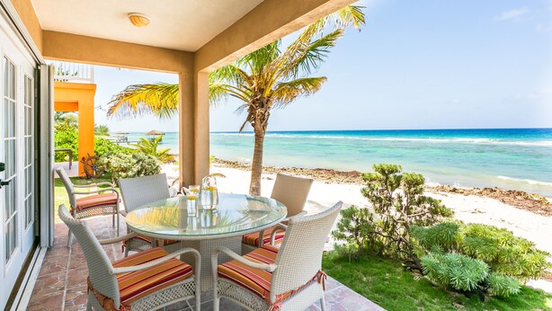 Mesa de vidrio con sillas de mimbre y cojines a rayas, con vista a una playa con agua azul cristalina y una palmera.