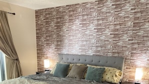 Un dormitorio con pared de diseño de piedra, cabecera capitonada, cama con ropa de cama estampada, dos lámparas de noche, un banco y alfombra con motivos.