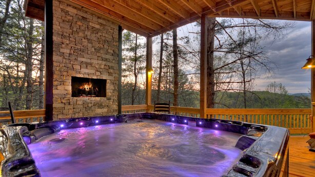 A hot tub with a stone wall and a fireplace, surrounded by a wooden deck and overlooking a forested area.