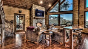 A log cabin interior with a stone fireplace, leather furniture, and large windows offering views of the outdoors.