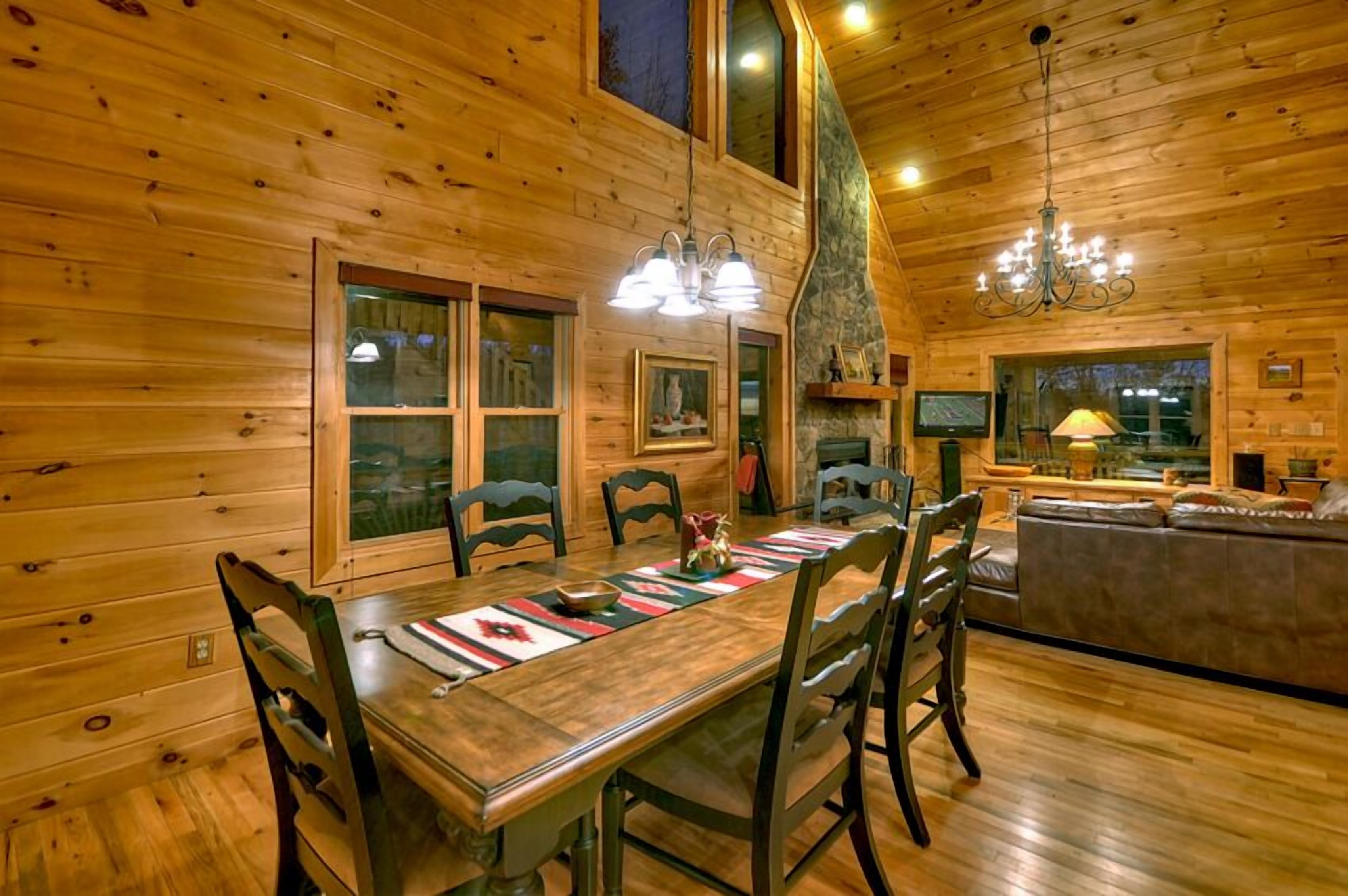 A wooden cabin interior with a dining table, chairs, a fireplace, and a living area with a sofa and TV.