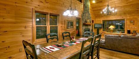 A wooden cabin interior with a dining table, chairs, a fireplace, and a living area with a sofa and TV.