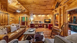 Interior acogedor de una cabaña con techo de madera, sofá de cuero, zona de comedor y televisor.