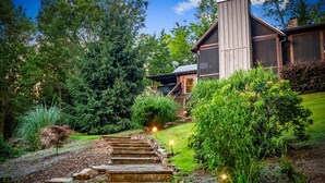 Una casa con una escalera de piedra que conduce a una terraza de madera, rodeada de exuberante follaje y árboles.