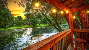 Una terraza de madera con vistas a un río y árboles, luces colgantes y un banco.