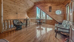 A wooden cabin with a sloped ceiling, a dining area with a table and chairs, a cushioned bench, and a view of trees through large windows.
