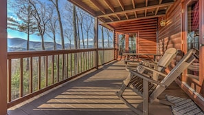 Una cabaña de madera con un porche cubierto, mobiliario exterior y vistas del bosque y las montañas circundantes.