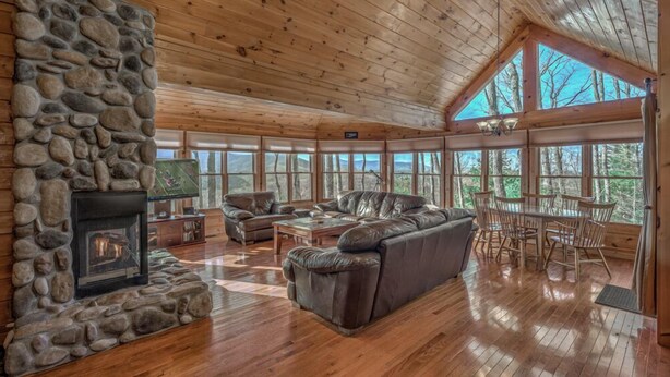 A spacious living room with a stone fireplace, leather sofa, wooden flooring, and large windows offering a view of the outdoors.