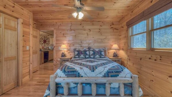 A wooden cabin bedroom with a bed, two bedside lamps, a ceiling fan, and a window.