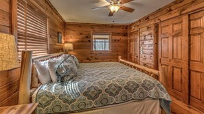 Une chambre dans une cabane en bois, avec un lit, une fenêtre, un ventilateur de plafond et des murs en bois.