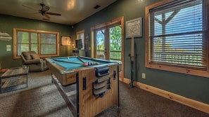 Une table de billard dans une pièce aux murs verts, avec un ventilateur au plafond et de grandes fenêtres équipées de stores.