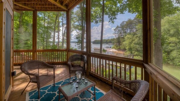 A covered porch with wicker furniture and a glass table, overlooking a lake and forested area. - Southern Comfort- Lake Front | 2 Level Dock | Paved Roads and Driveway (Morganton)