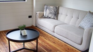 Una habitación con un sofá de tonos claros, una mesa de centro redonda negra con una planta pequeña, suelo de madera y una ventana con persianas.