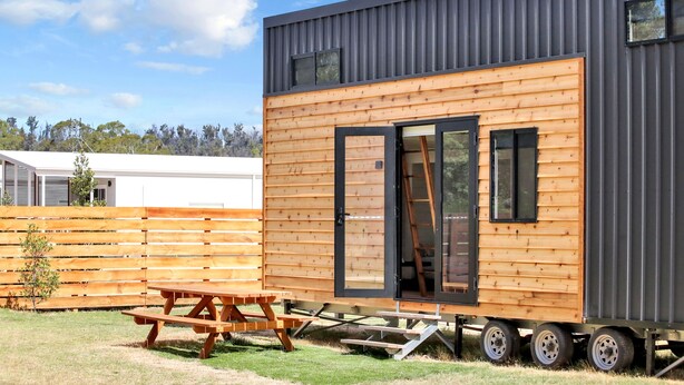 Una casa móvil moderna con revestimiento de madera y techo gris, que cuenta con una puerta corredera y un ventanal pequeño.